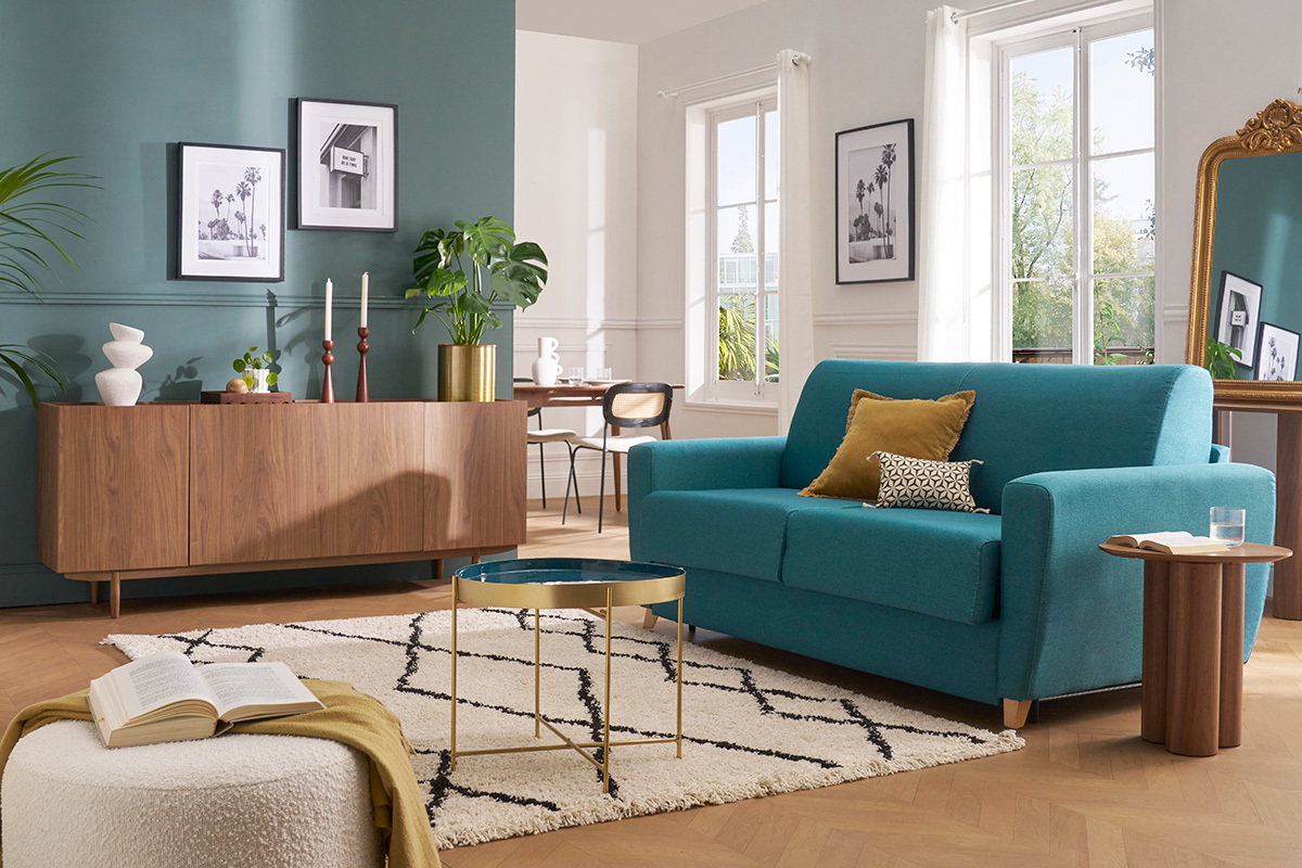 Canap� convertible bleu canard dans un salon lumineux avec table d'appoint en bois.